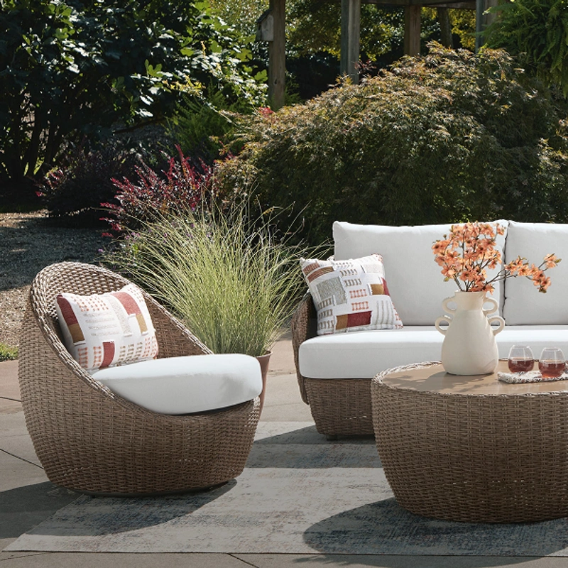 Wicker outdoor furniture set with white cushions and colorful pillows including a central table with two drinks and a white vase with orange flowers